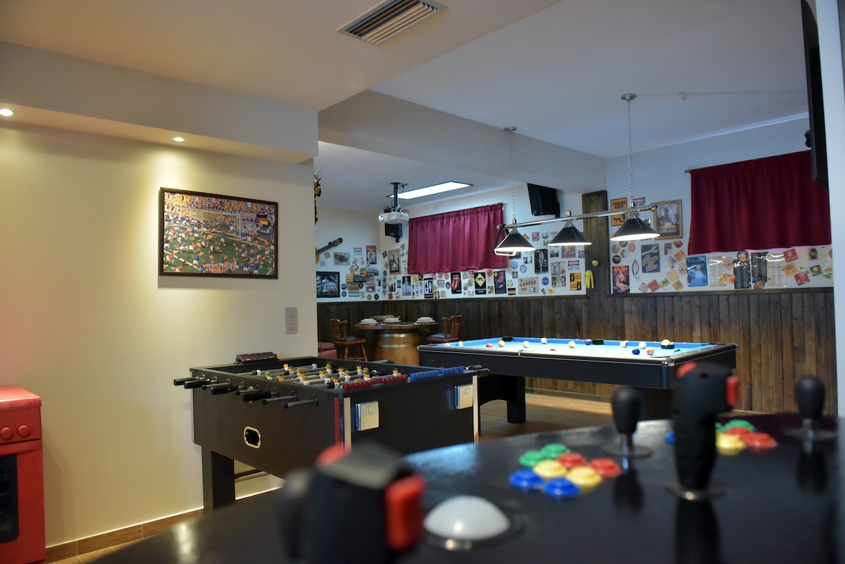 The spacious entertainment area features a foosball table, a pool table, and a collection of wall art and photographs. Warm lighting is provided by recessed fixtures, and a large window allows natural light to flow in. Comfortable seating areas are visible in the background.