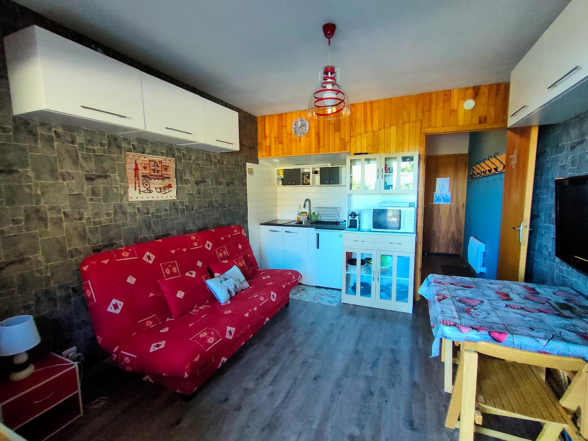 A living area features a red sofa bed and two tables, one with a patterned tablecloth. The kitchen area is equipped with white cabinets, an oven, and a microwave. Natural light enters through a nearby doorway, highlighting the stone-accented wall.
