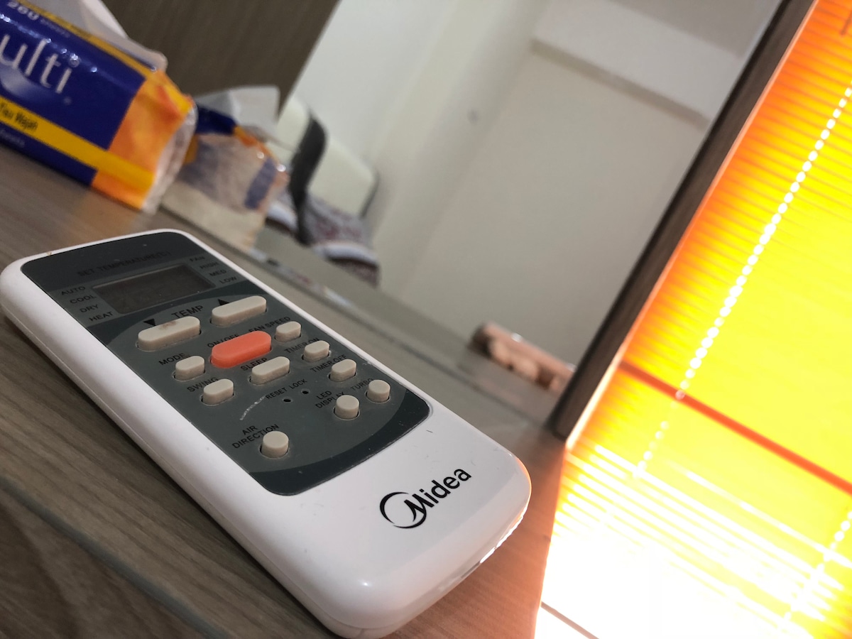 A remote control rests on a wooden surface, featuring buttons for various functions. In the reflection, a partially visible room contains soft lighting from a window covered with blinds, creating a warm ambiance. Items are neatly arranged in the background.
