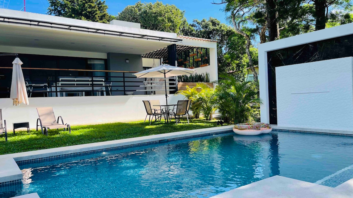 A private pool area features inviting blue water, surrounded by a green lawn. A shaded dining set with chairs and an umbrella is positioned nearby, alongside two lounge chairs. The villa’s modern structure is visible in the background, framed by lush trees and greenery.