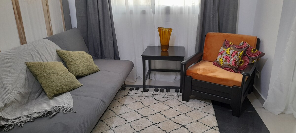 A comfortable seating area features a gray sofa with decorative pillows and an orange-accented chair. A small black table sits between them, with a yellow vase adding a vibrant touch. Soft lighting filters through sheer curtains, creating a cozy ambiance.