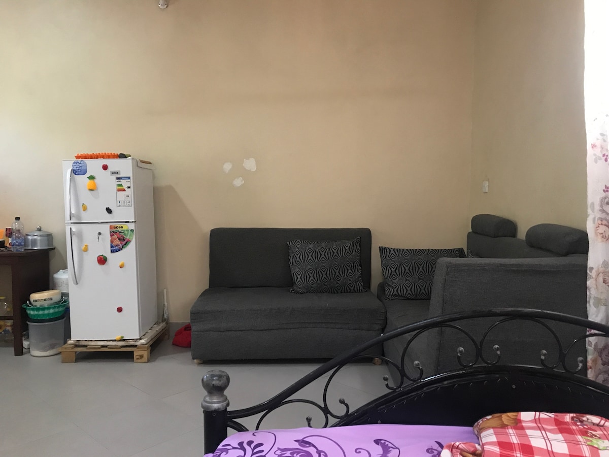 A living area features a grey fabric sofa set with patterned cushions, positioned near a small dining table. A white refrigerator is seen to the left, with various items placed on top. The walls are painted in a light tone, complementing the tiled floor.