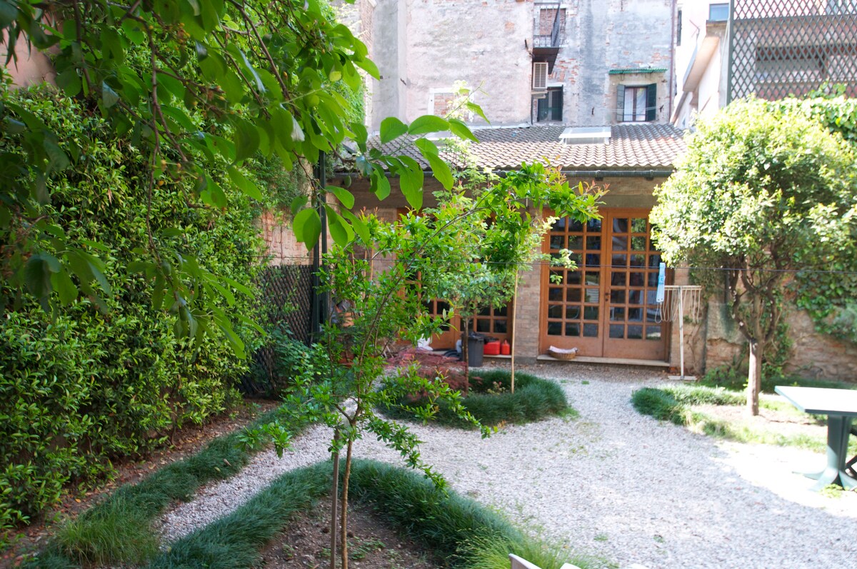 A serene garden space is framed by lush greenery, featuring a mix of mature shrubs and small trees. A gravel pathway meanders through the garden, leading to a building with glass doors that bring natural light to the area. The atmosphere suggests a peaceful retreat.