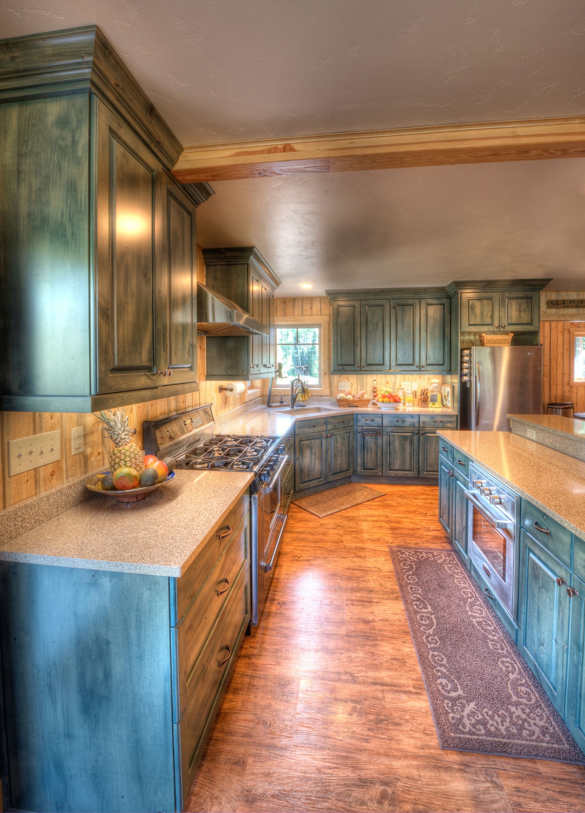 The kitchen features rich blue-green cabinetry complemented by wood accents. Stainless steel appliances and a large countertop enhance functionality. A bowl of fresh fruit sits on the counter, while warm lighting creates an inviting work area. The attractive wood flooring adds to the rustic charm.