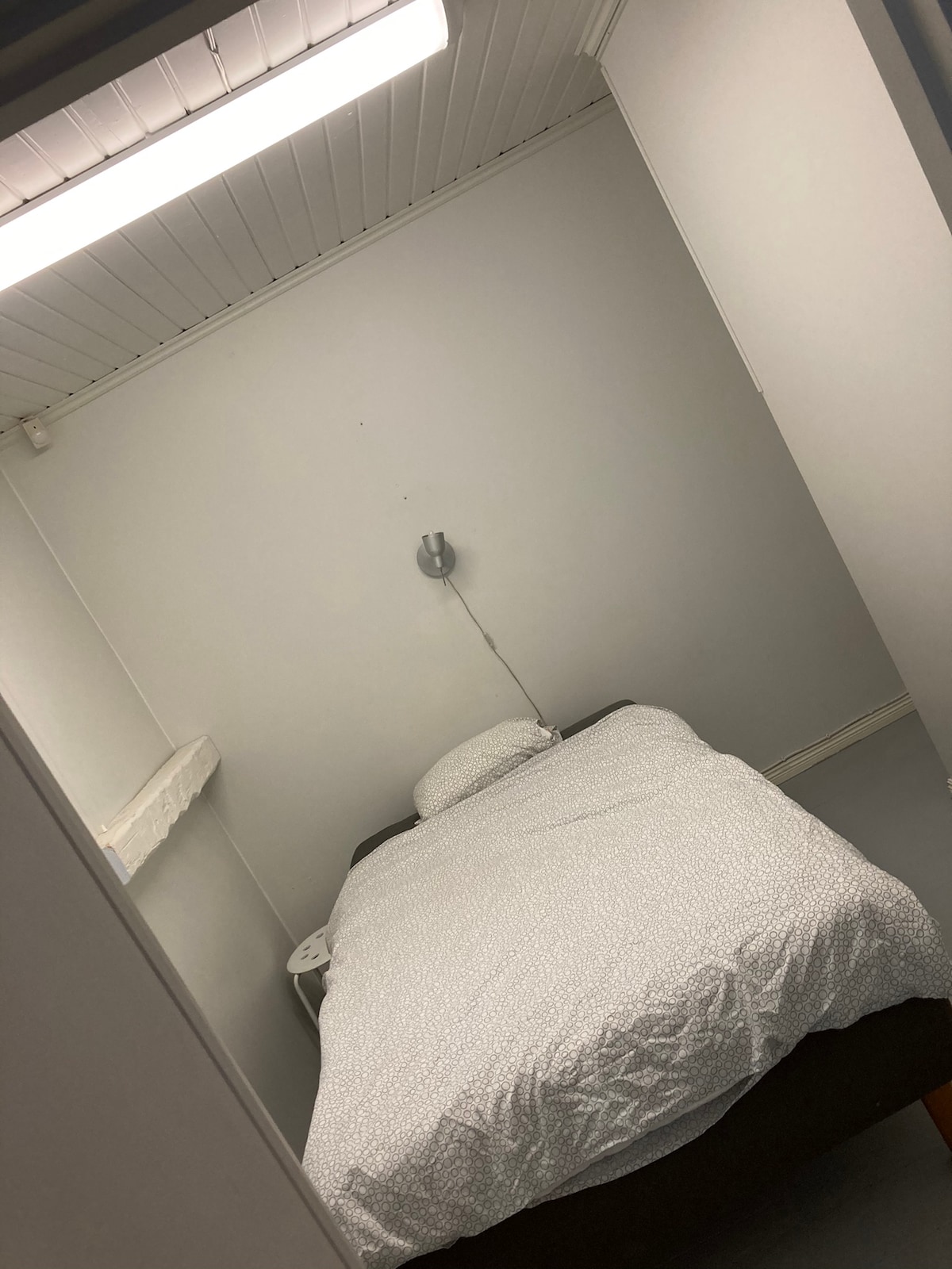 A modest sleeping area is presented with a single bed dressed in light bedding. A wall-mounted reading light offers soft illumination. The walls are painted in a neutral tone, contributing to a clean, simple aesthetic.