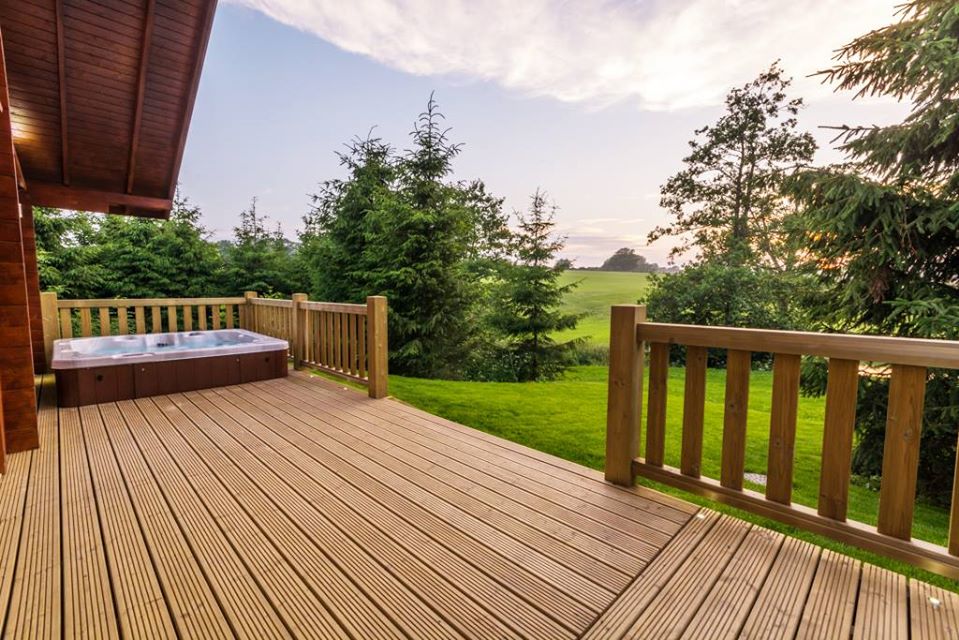 A wooden deck extends outwards, featuring a private hot tub set against a backdrop of lush greenery. The tranquil landscape is framed by tall trees, while soft evening light casts a gentle glow over the scene.