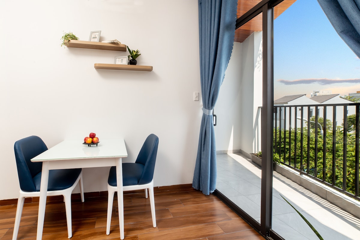 A dining area features a small white table with a bowl of fresh fruit. Two blue chairs are placed around the table. Floor-to-ceiling glass doors open to a spacious balcony, allowing natural light to fill the room. Soft blue curtains frame the window.