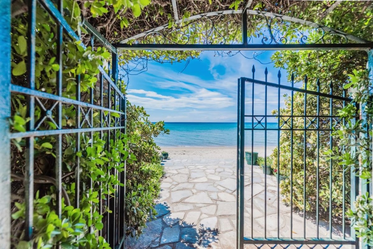 A wrought iron gate is visible, partially opened to reveal a clear view of a sandy beach and the calm, blue sea beyond. Green foliage frames the entrance, enhancing the natural setting.