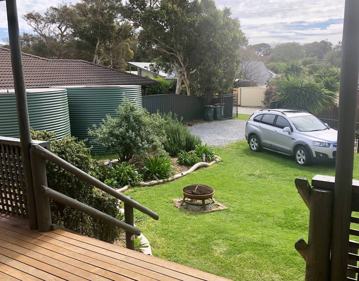 A grassy outdoor area is visible, featuring a circular fire pit surrounded by small stones. Various plants and shrubs are scattered throughout the yard, with a silver vehicle parked nearby. A garden is to the side, and a gravel pathway leads to the entrance.