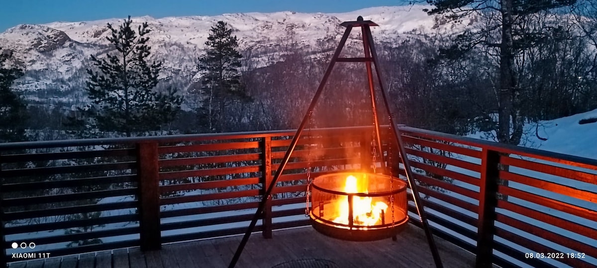 A fire pit is suspended on a deck, providing warmth and light against the backdrop of snow-covered mountains. The surrounding trees add to the natural setting, while the evening sky casts a peaceful ambiance over the scene.