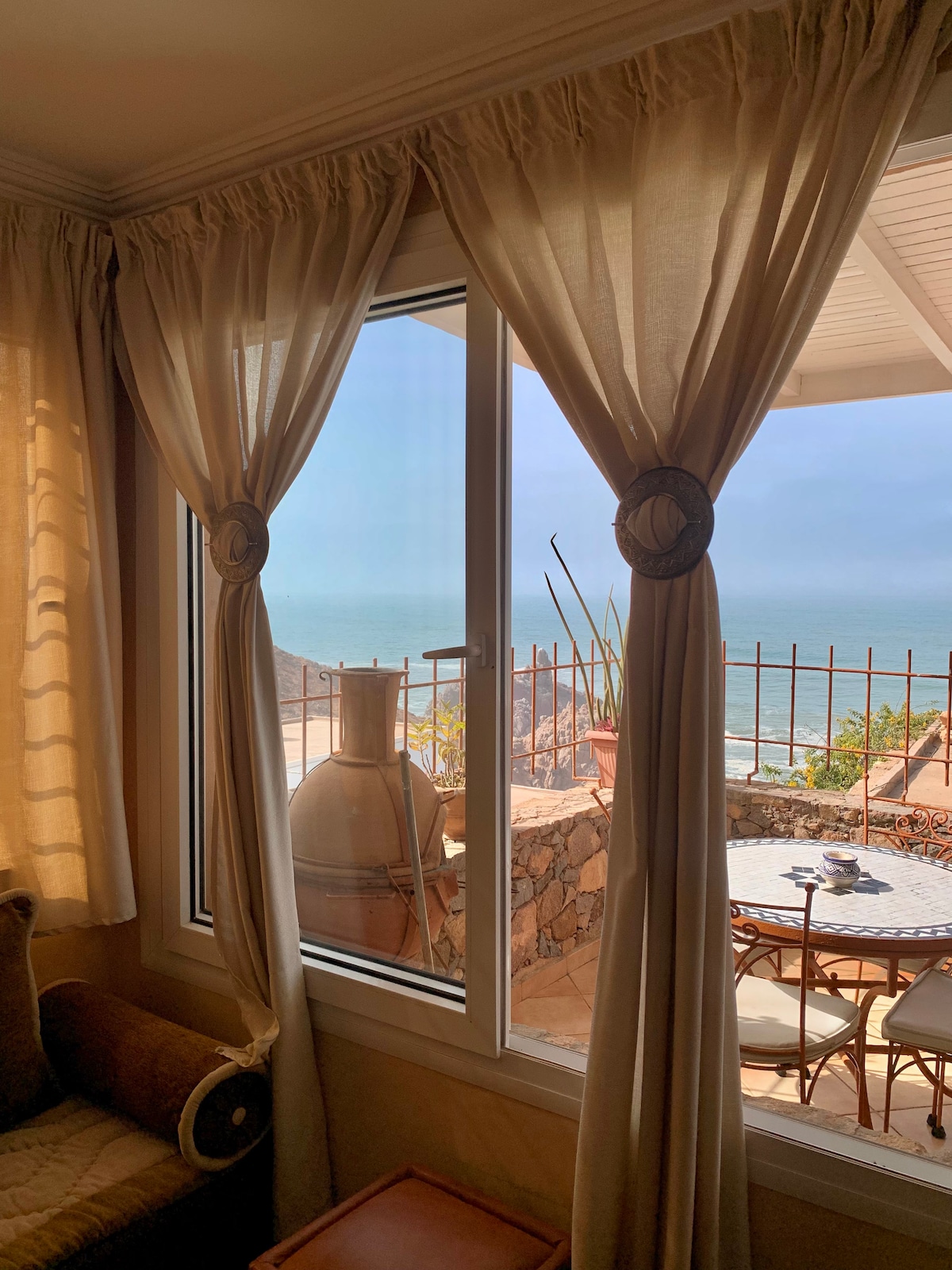 A large window frames a coastal view, with sheer curtains gently draping to the sides. Outside, a small terrace is visible, featuring a round table and chairs. The natural light illuminates the interior, enhancing the cozy atmosphere.