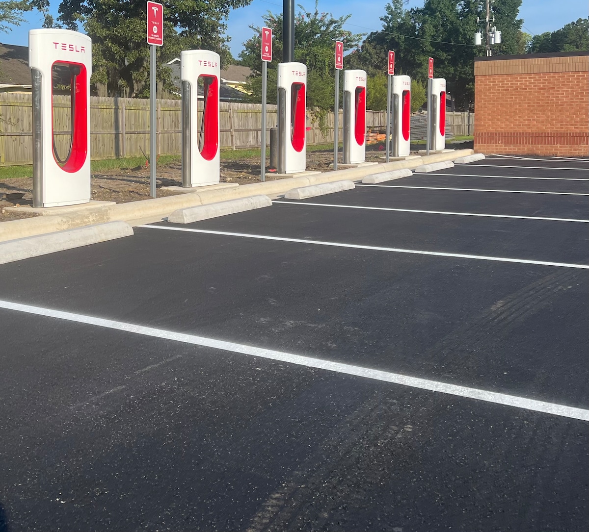 A row of Tesla Supercharging stations is displayed in a well-maintained parking area. Each station features a white and red design, with the Tesla logo prominently shown. The spaces are lined with fresh white markings on a newly paved surface, surrounded by a low wooden fence and greenery.