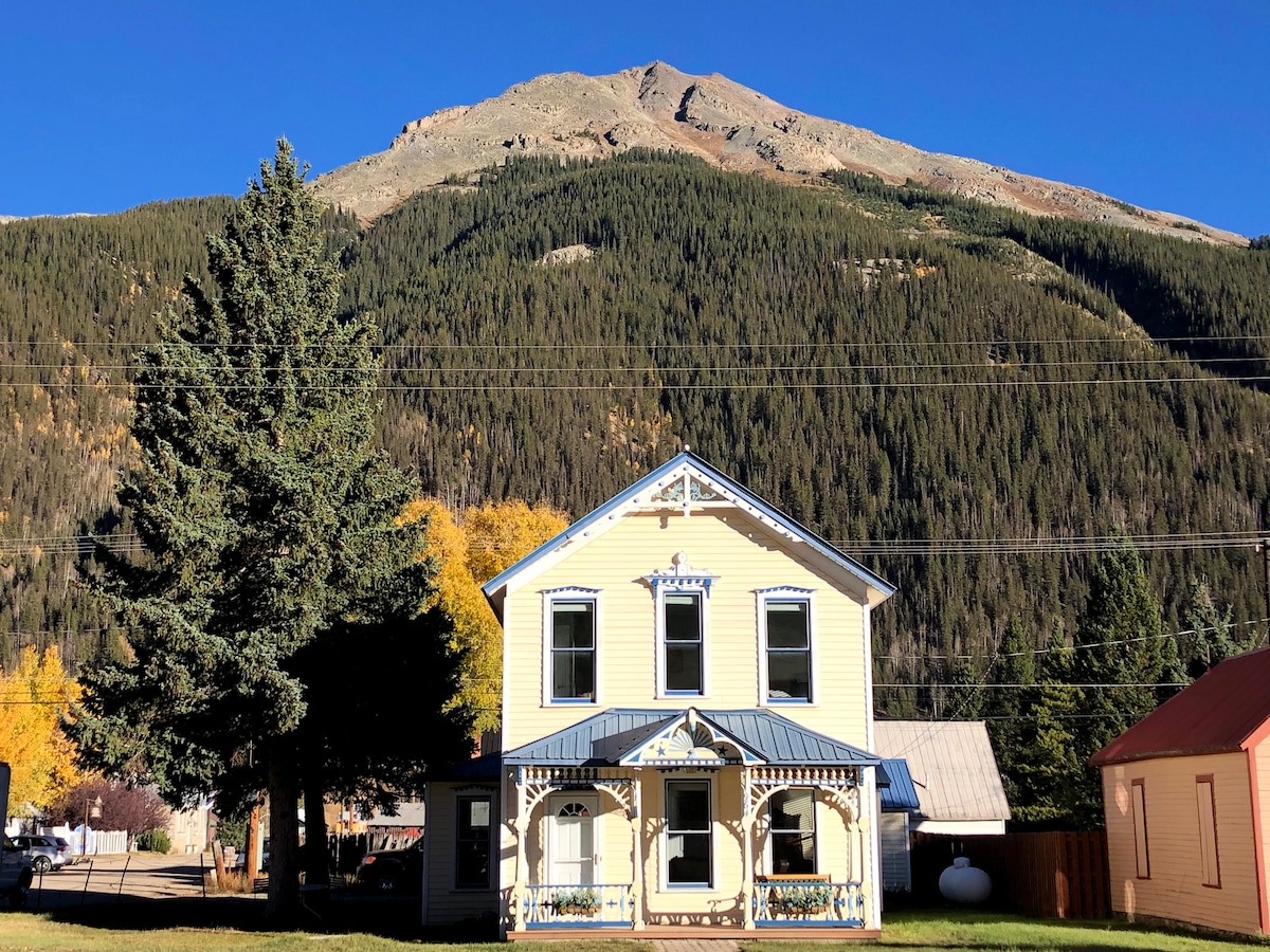 A historic Victorian home is set against a backdrop of lush green mountains. The two-story structure features a welcoming front porch and a vibrant facade. Tall trees add to the charm, while the clear blue sky enhances the serene environment.