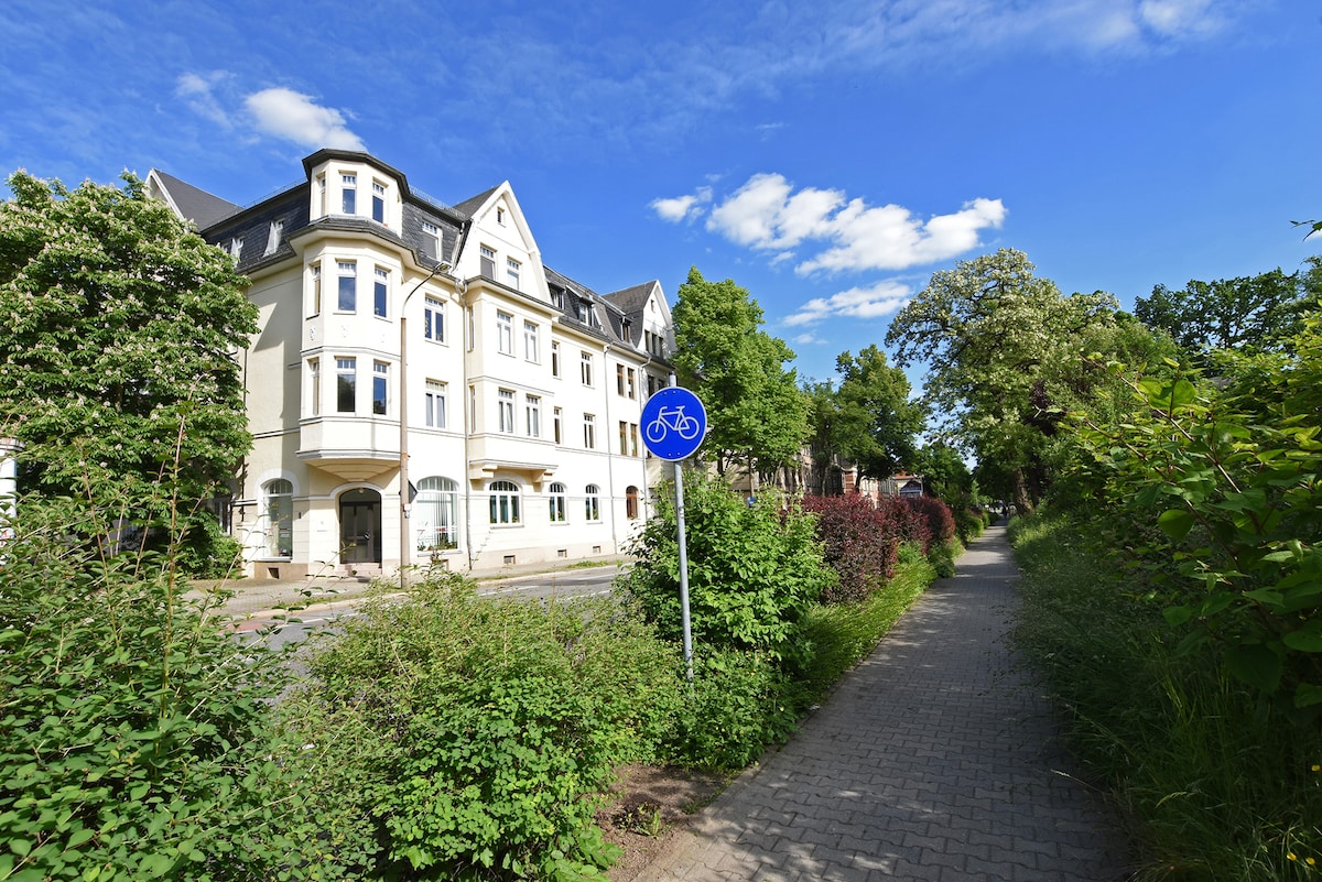 An inviting pathway is lined with greenery, leading past a well-maintained building featuring an elegant facade. A blue bicycle sign is visible, indicating a bike-friendly area. The sky is bright with a few scattered clouds, contributing to the serene atmosphere of the surrounding neighborhood.