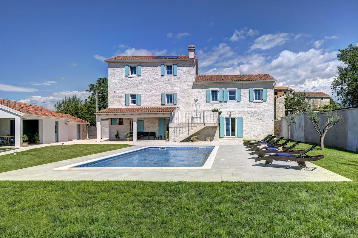 A spacious villa exterior is showcased, featuring a bright stone façade and blue-shuttered windows. A large rectangular saltwater pool is set on a deck surrounded by sun loungers, with a green lawn and landscaped garden enhanced by clear skies.