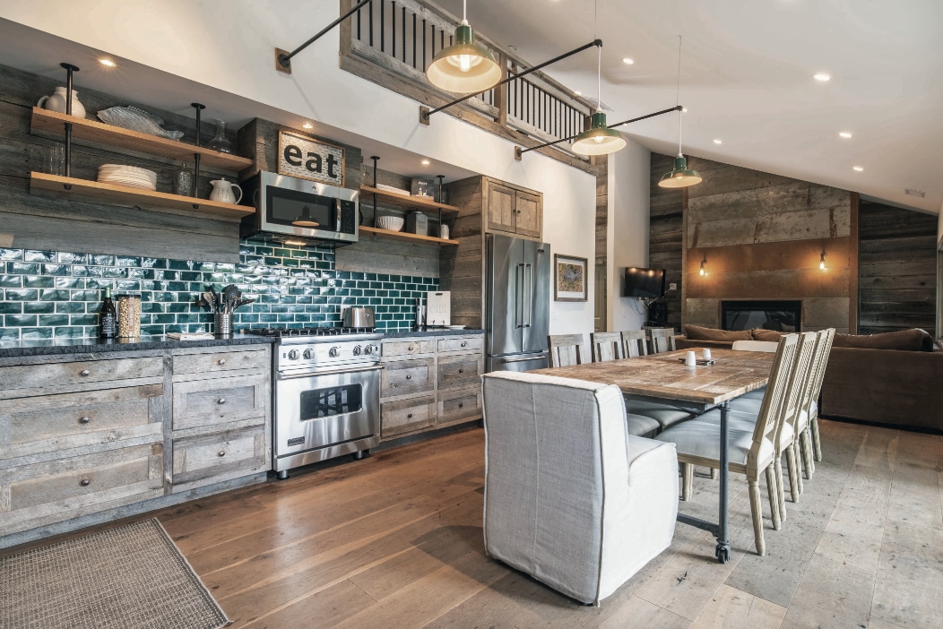 A spacious gourmet kitchen features rustic wooden cabinetry and modern stainless-steel appliances. A large dining table with seating is positioned nearby. A decorative sign with the word 'eat' adds character, while backsplash tiles in a deep turquoise provide a vibrant contrast to the neutral tones.