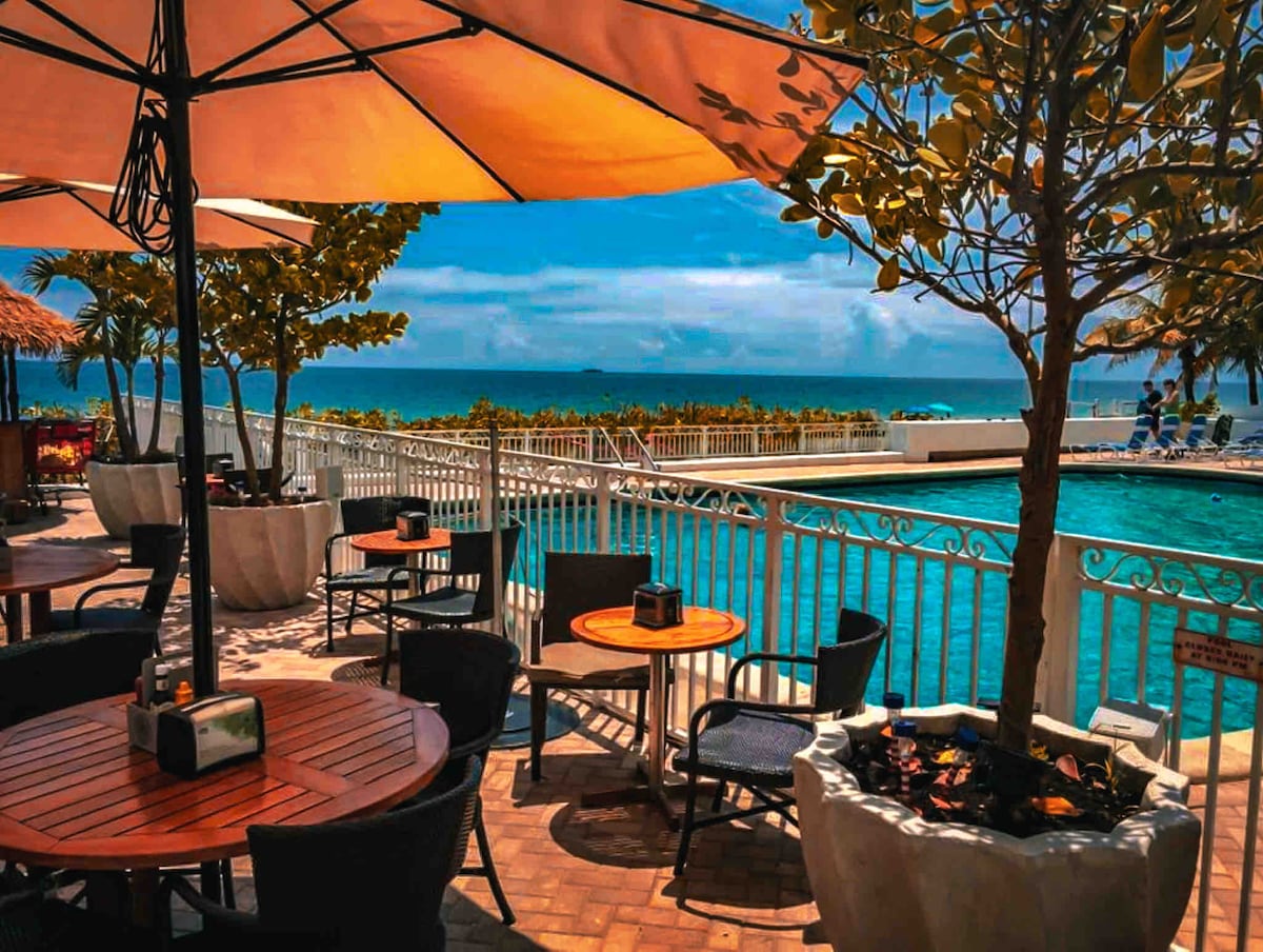 An outdoor seating area features multiple round wooden tables surrounded by dark chairs. Shade is provided by large umbrellas, while vibrant greenery is present in pots nearby. The swimming pool is visible in the background, along with a glimpse of the ocean beyond.