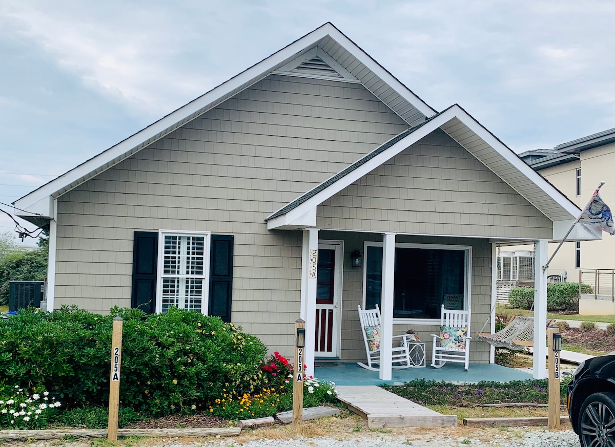 The exterior of the cottage showcases a light gray shingle facade with a welcoming front porch. Two rocking chairs are positioned on the porch, surrounded by blooming flowers and greenery. A gravel pathway leads to the entrance, adding to the coastal charm.
