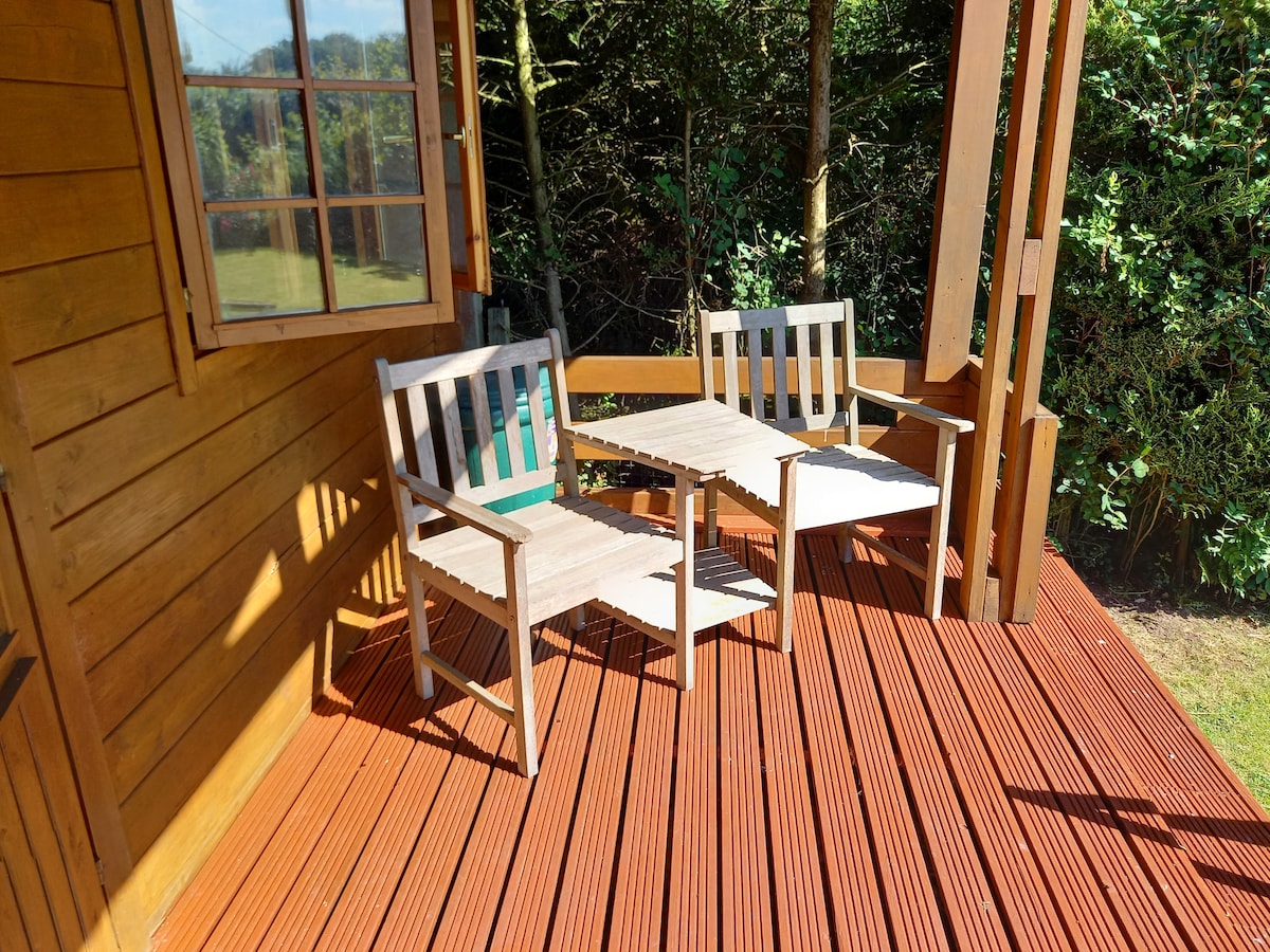 A small outdoor seating area features two wooden chairs and a matching table, set on a wooden deck. The lush greenery of the surrounding garden is visible in the background, offering a calm and private setting.