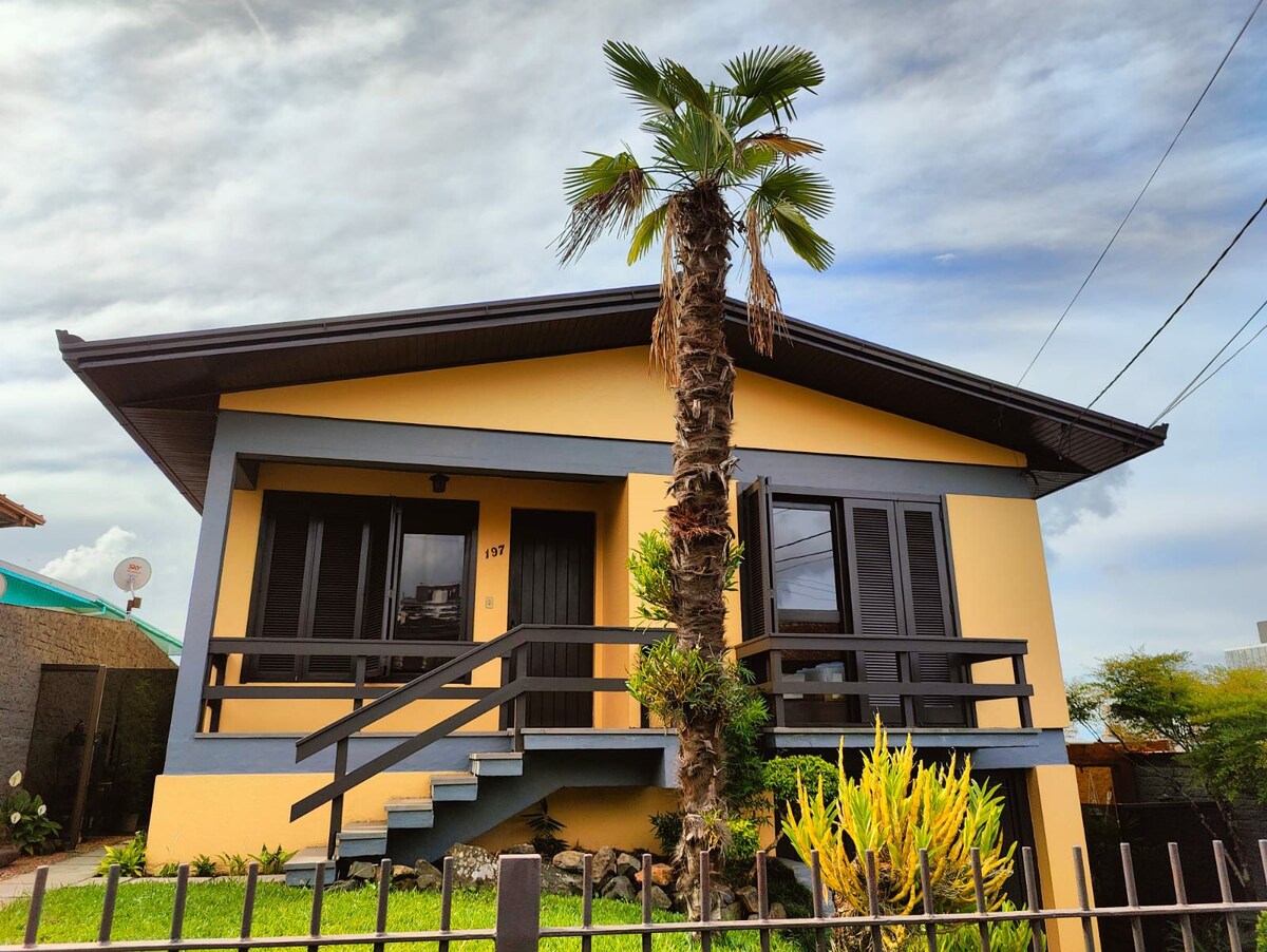 The exterior of a yellow and black house is visible, featuring a front staircase and a wooden railing. A palm tree stands in the yard, complemented by vibrant green plants. The structure includes large windows that offer a welcoming appearance.
