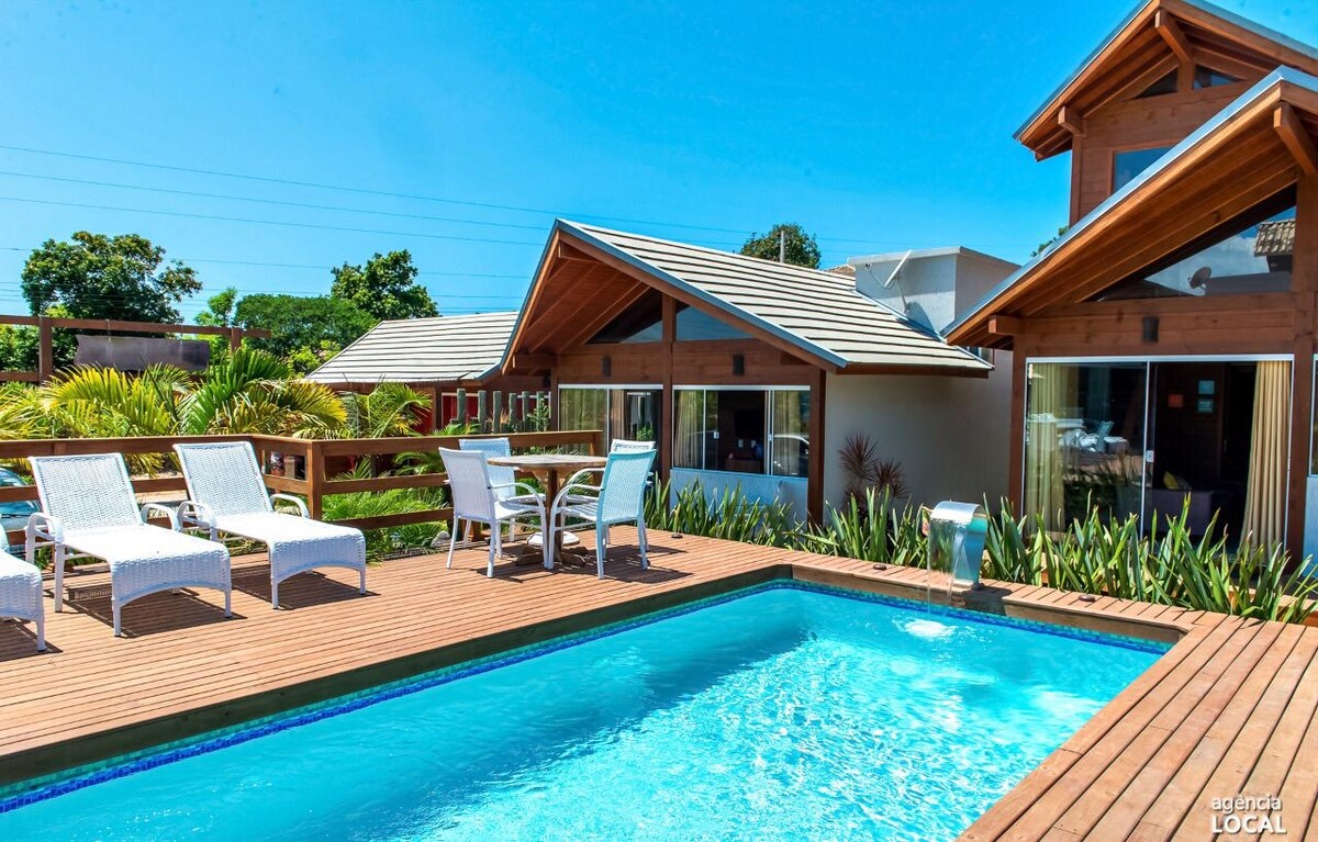 A wooden deck surrounds a clear swimming pool, with four lounge chairs positioned nearby. The house features large glass windows and a sloped roof, allowing natural light into the interior. Lush greenery borders the area, enhancing the serene outdoor space.