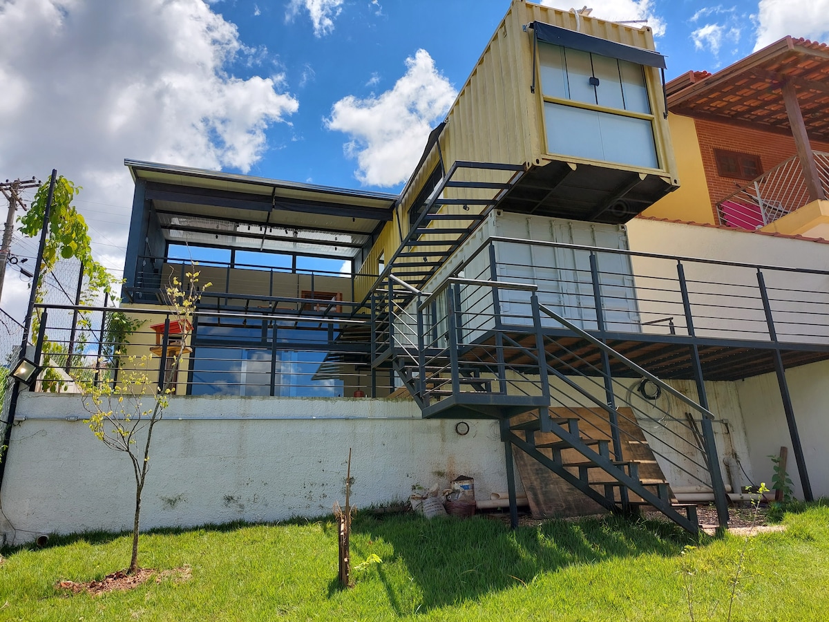 A modern container home is showcased with a multi-level design. A staircase leads to a spacious deck, with large glass windows reflecting the surrounding greenery. The exterior features a blend of natural and industrial materials, creating a visually striking facade.