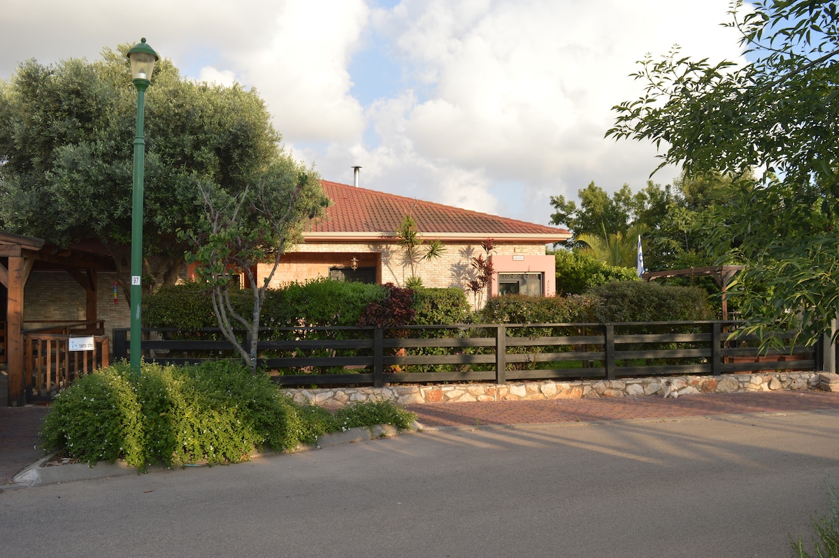 The villa's exterior features a charming brick facade surrounded by well-maintained greenery. A wooden fence borders the property, adding privacy. The roof is red, and trees provide shade. Soft clouds are seen in the background, enhancing the serene atmosphere.