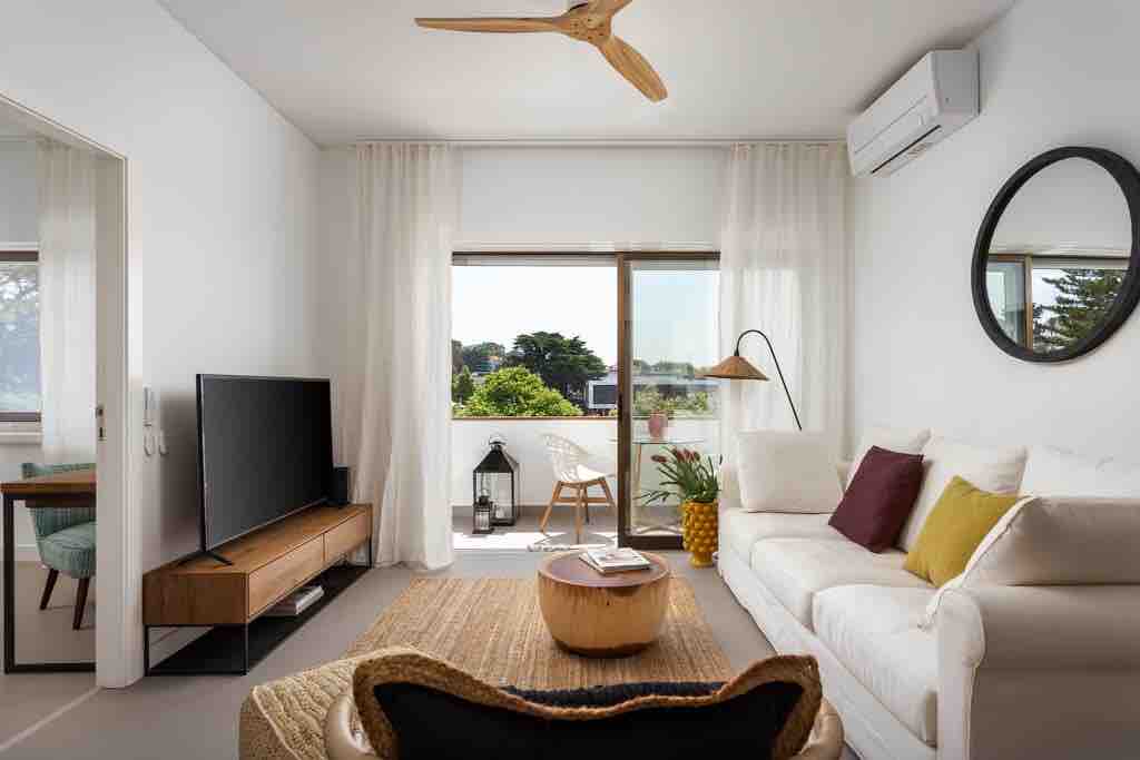 A spacious living room is highlighted by a large window with a view of greenery. A comfortable sofa with accent pillows is paired with a modern coffee table. A flat-screen TV is mounted on a wooden console, while a floor lamp and decorative elements enhance the space.