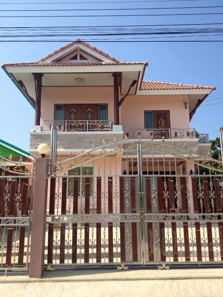 A two-story house is depicted with a pink exterior and a traditional roof. The structure is fronted by a decorative metal gate and complemented by a spacious porch. Clear skies are visible above, enhancing the overall appearance of the residence.