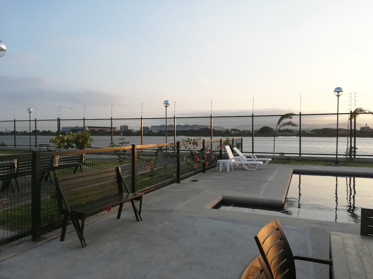An outdoor area features a pool adjacent to the water, with poolside loungers and benches arranged for seating. Foliage and flowers add greenery along the perimeter, while lamps provide illumination as the sun sets in the background.