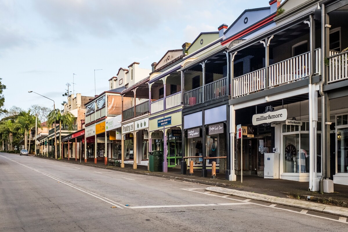 A row of charming storefronts lines the quiet street, featuring a variety of colors and architectural styles. Covered verandas and decorative railings add character, while the gentle morning light casts a soft glow over the scene, inviting a leisurely exploration of local shops and cafes.