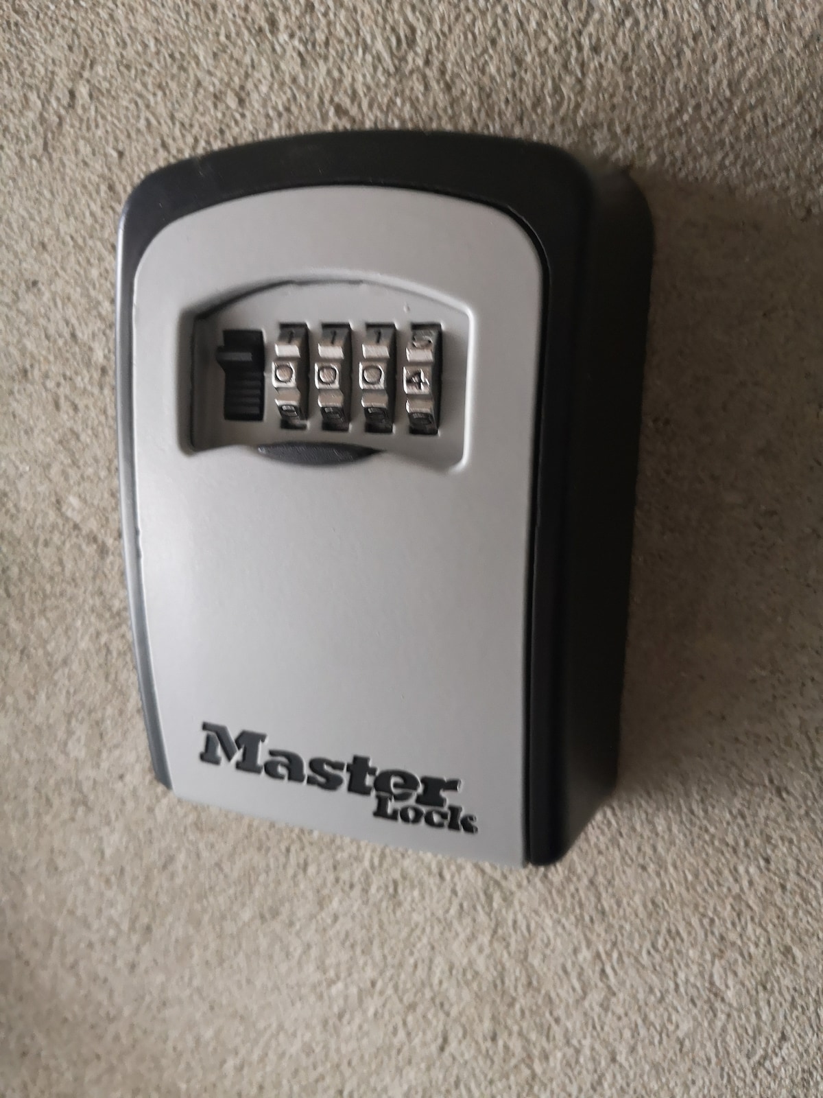 A secure keybox is mounted on a light-toned wall, featuring a four-digit combination lock. The brand name 'Master Lock' is clearly visible on the front, providing a robust and practical access solution.