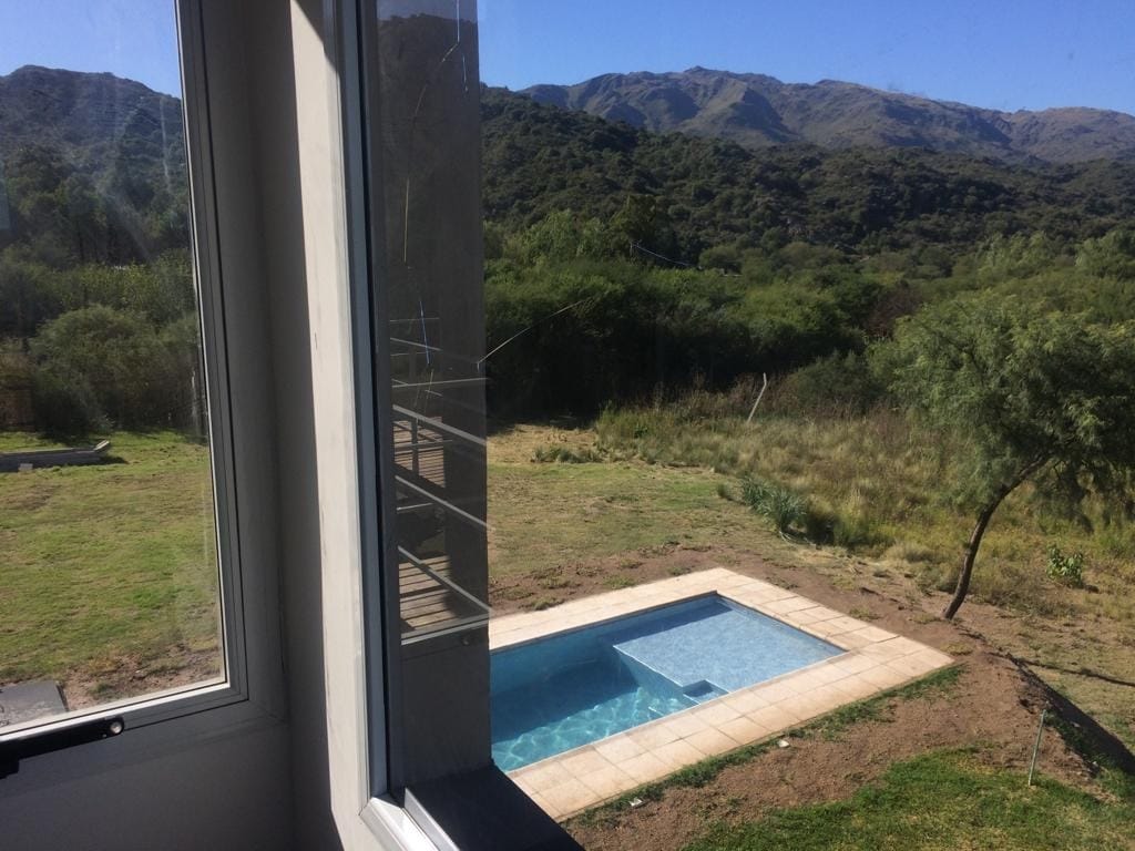 A view from a large window showcases a private pool set within a grassy outdoor space. The surrounding landscape features hills and greenery, providing a serene backdrop. Sunlight reflects off the water, enhancing the tranquil atmosphere.