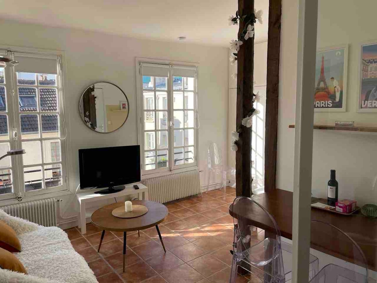 A bright living area is visible, featuring exposed beams and large windows that allow natural light to fill the space. A circular mirror reflects the room, while a coffee table sits in front of a cozy sofa. A television and dining area with clear chairs are also present.