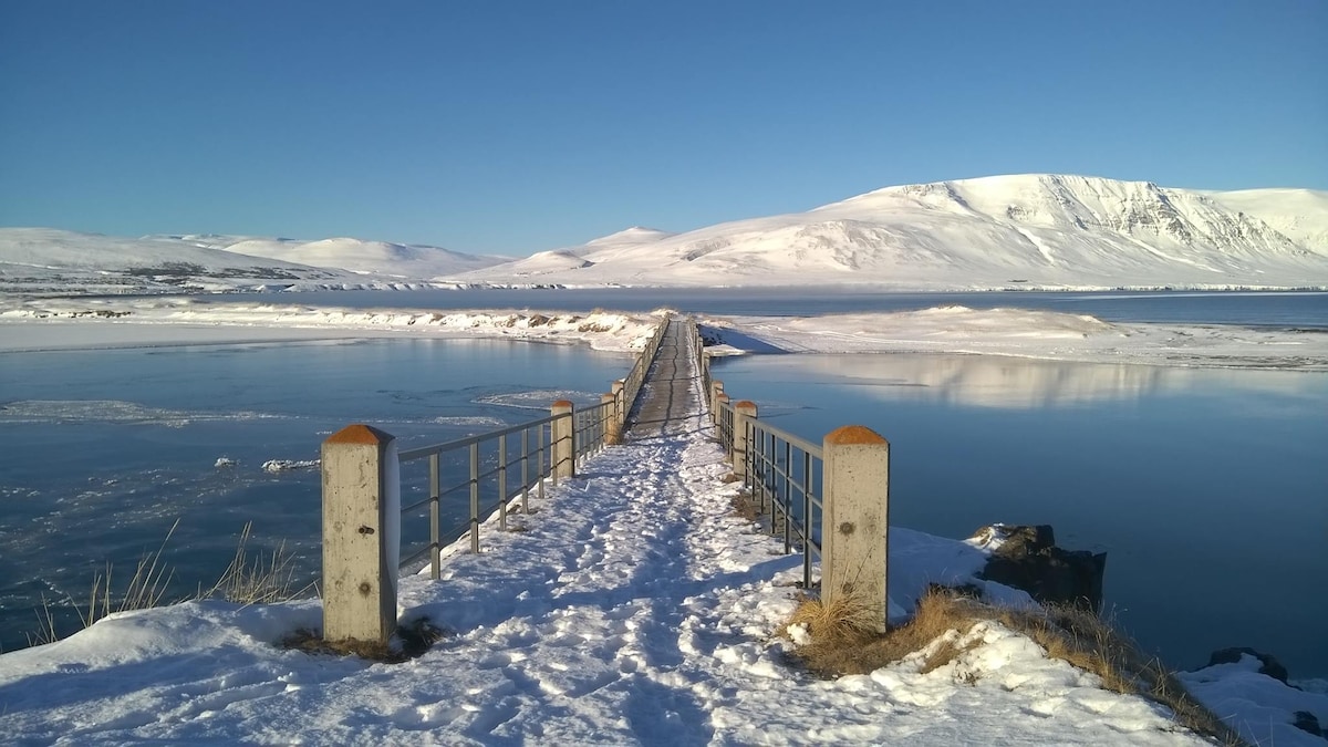 A snow-covered pathway leads across a bridge, with stone pillars at each end marking the entry. The serene water reflects a clear blue sky, while distant snow-capped mountains frame the horizon, showcasing the peaceful beauty of the natural landscape.