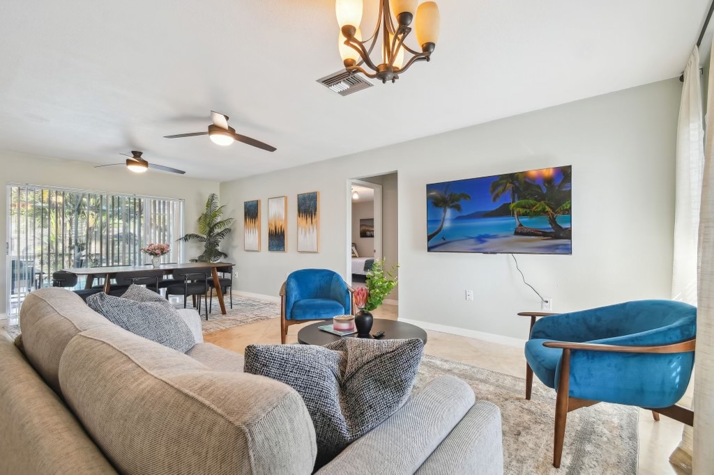 A bright living area is highlighted by a large sectional sofa and two blue accent chairs. A mounted flat-screen TV displays a serene beach scene. A dining area with a long table is visible in the background, enhancing the inviting layout.