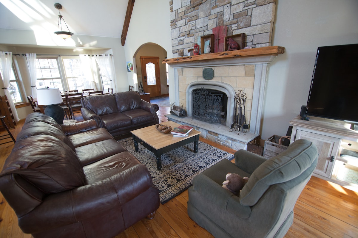 An inviting living space features a large stone fireplace as a focal point, surrounded by comfortable leather seating. Natural light floods the room through windows draped in sheer curtains, while a central coffee table rests on a decorative rug, adding warmth and character.