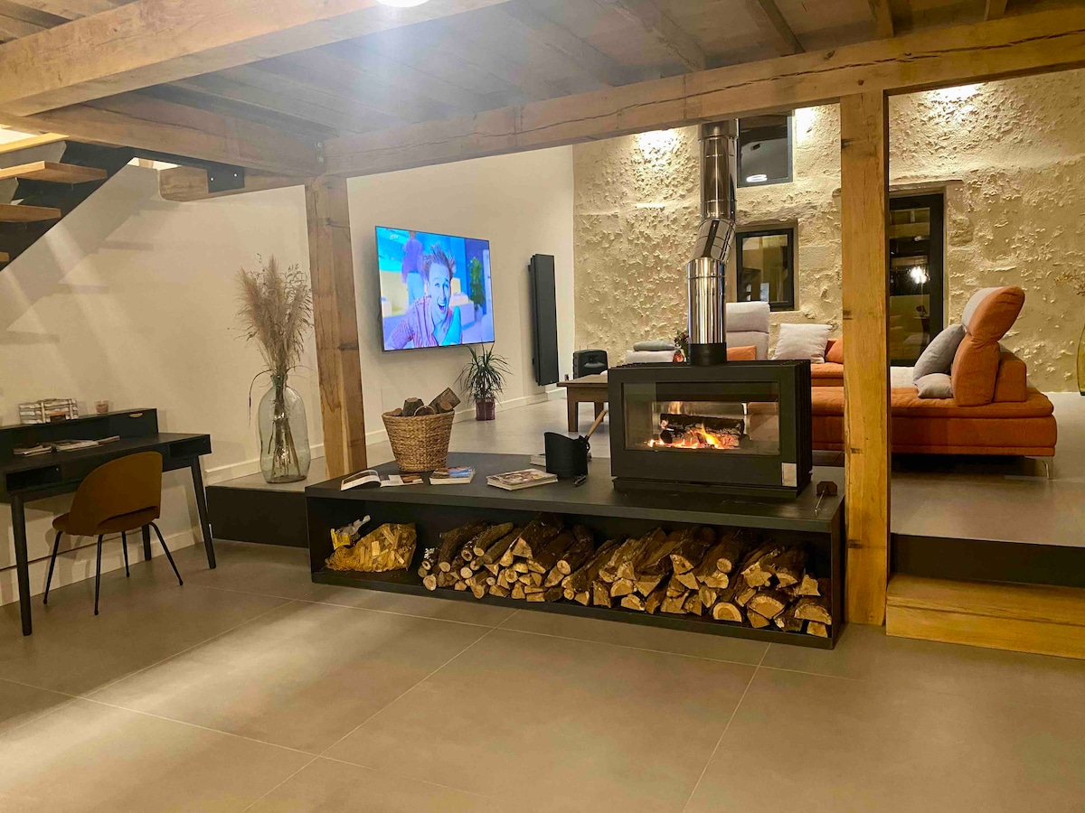 A spacious living area is featured, showcasing a cozy seating arrangement with a large television mounted on the wall. A modern fireplace sits centrally, flanked by neatly stacked firewood. Natural elements are included with decorative plants, and muted tones enhance the contemporary décor.