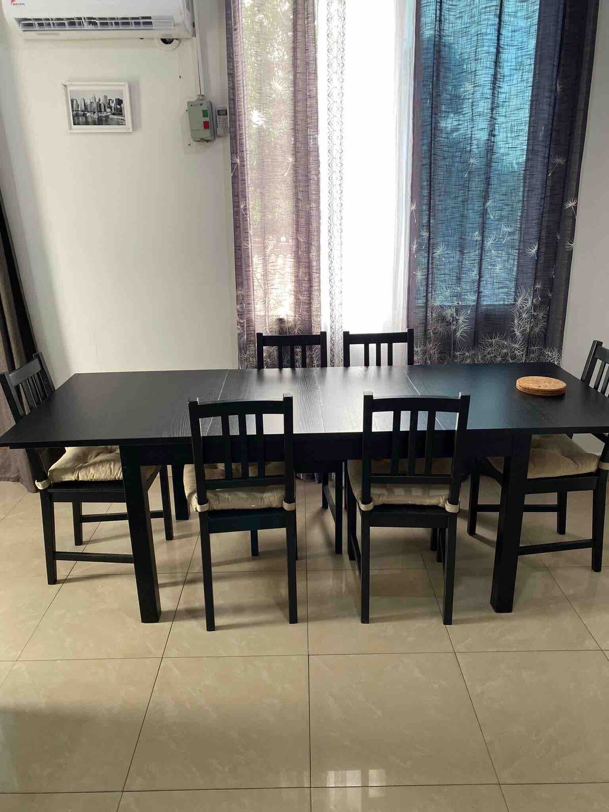 A dining area features a rectangular black table surrounded by six matching chairs. Natural light filters through sheer curtains, illuminating the space. A decorative bowl sits on the table, adding a subtle touch to the minimalist design.