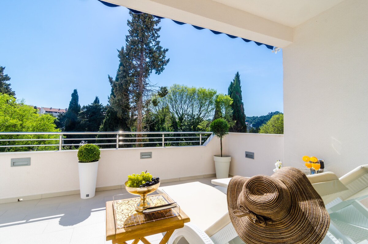 A furnished balcony is visible, featuring a wooden table with a fruit platter and two drinks. Sun loungers are positioned nearby, with a wide view of a lush garden and trees. Clear blue skies frame the scene, enhancing the outdoor space's tranquility.
