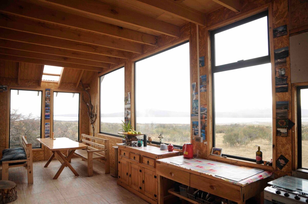 A bright and airy common area features large windows that provide expansive views of the surrounding landscape. A wooden dining table sits in the center, accompanied by benches. Various wall decorations enhance the rustic charm of the space, while a countertop displays kitchen essentials.