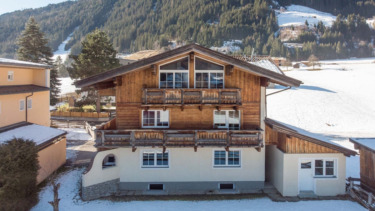 The exterior of the alpine-style lodge is visible, featuring a combination of wooden and stone accents. Snow-covered ground extends into the backdrop of rolling green hills and mountains, while multiple balconies provide a scenic view. A pathway leads up to the entrance, framed by evergreen trees.