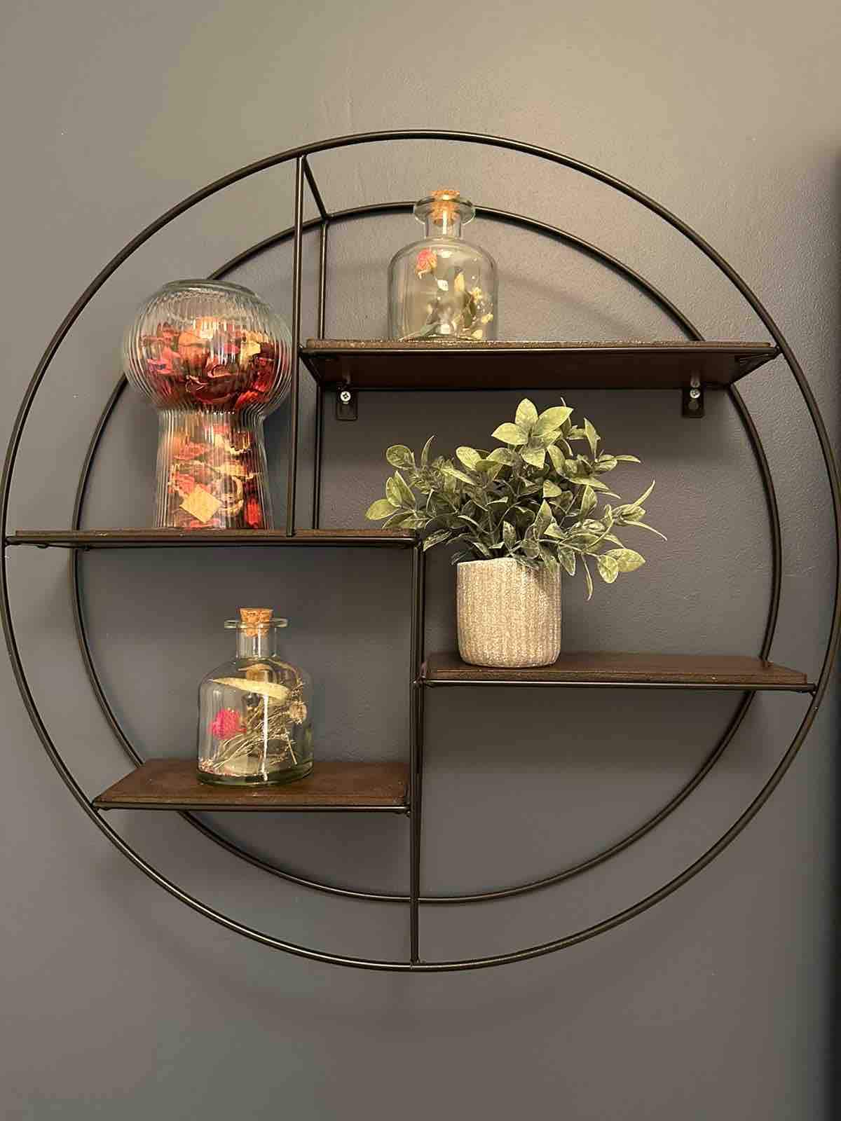 A circular wall shelf is showcased against a dark background. It features multiple wooden shelves holding decorative glass containers filled with colorful items and a potted plant, adding a touch of greenery and style to the space.