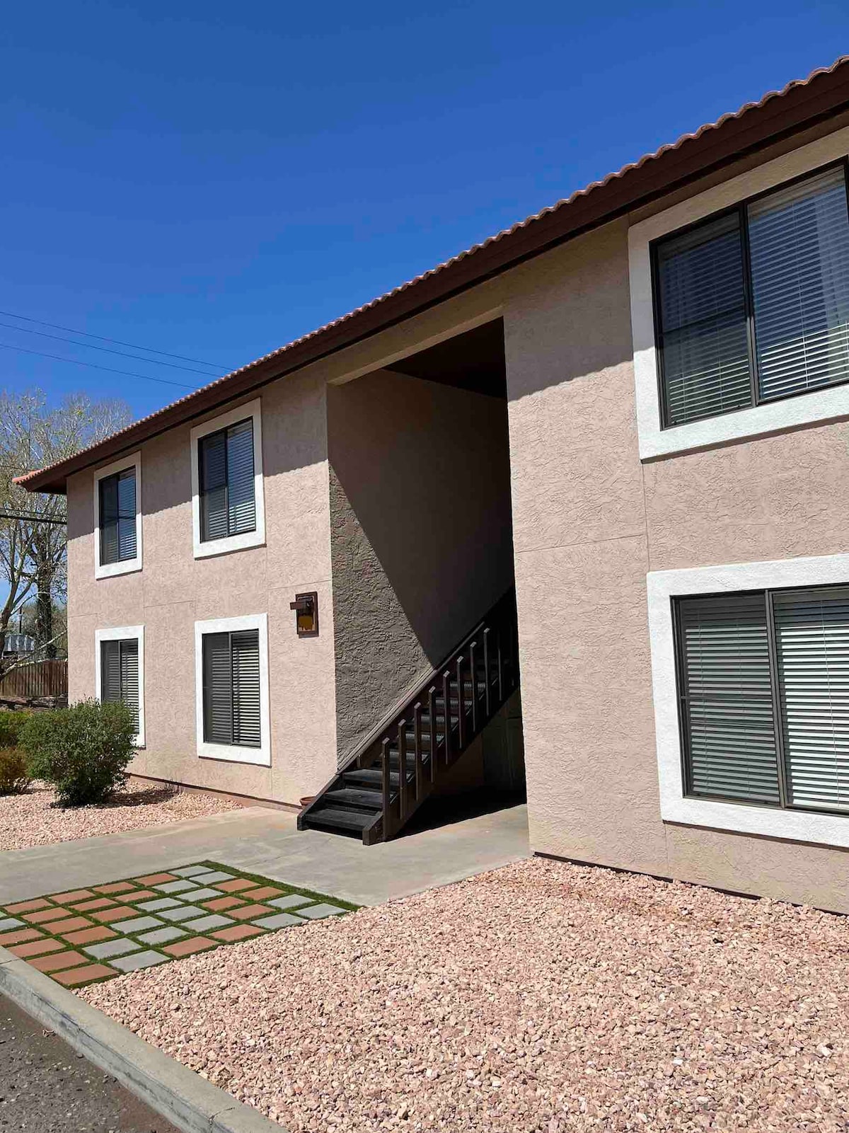 The exterior of a two-story apartment building is visible, featuring a neutral-toned facade. Windows are framed in white, allowing natural light to filter in. A set of stairs leads to the entrance, with a decorative pathway in the foreground and landscaping of small shrubs and gravel.