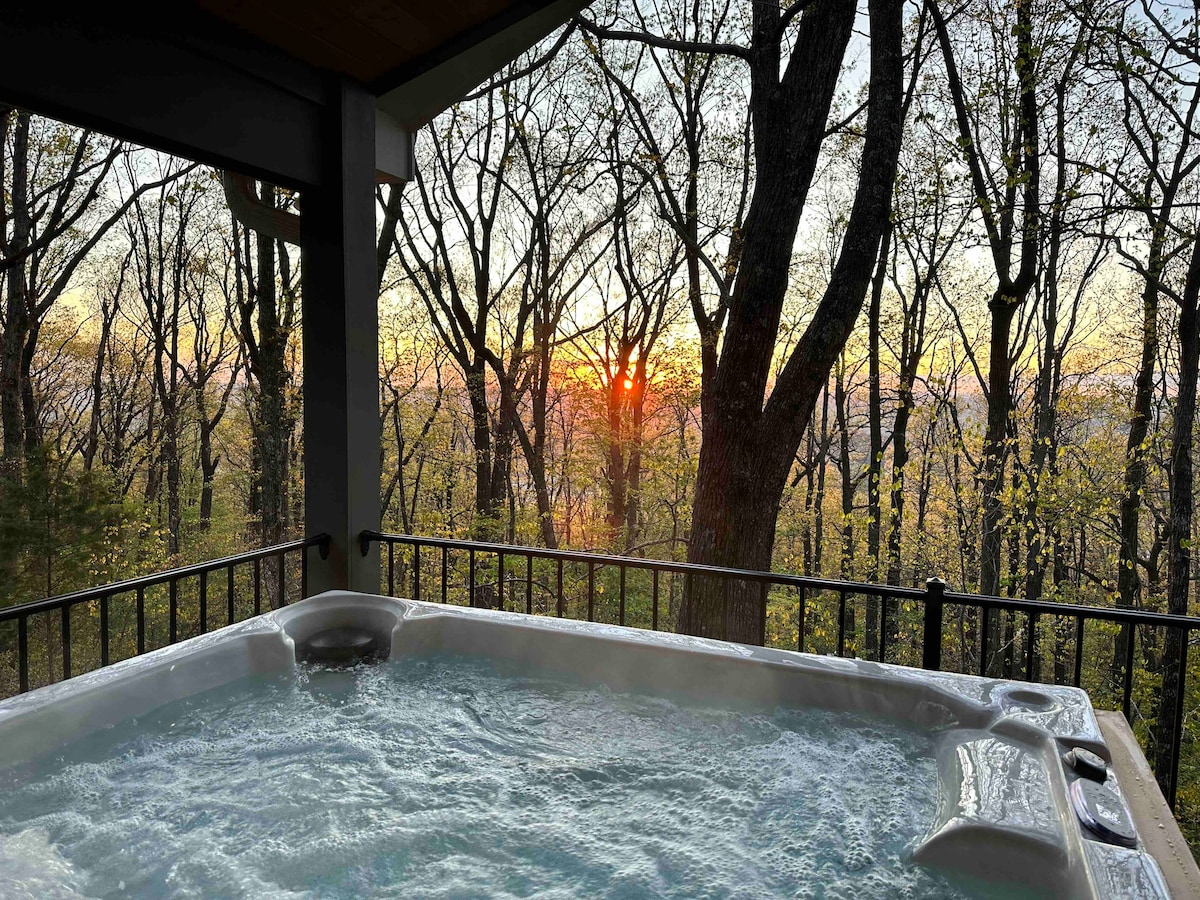 A hot tub is situated on a covered porch, overlooking a tranquil forest setting. Trees frame the view, while a sunset casts warm tones across the landscape. The bubbling water reflects the serene atmosphere of the surrounding nature.