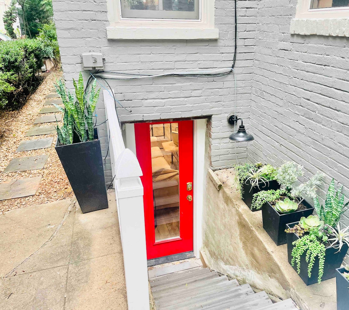 The entrance to the studio is depicted, featuring a vibrant red door framed by white trim. Concrete steps lead downward, flanked by potted plants with lush green foliage. The building's brick exterior is painted gray, creating a contrast with the colorful door.