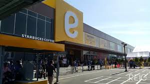 A modern commercial building is showcased with prominent yellow signage featuring the letter 'e.' The Starbucks Coffee sign is visible at the entrance, while a line of customers can be seen outside, indicating a popular gathering spot. Bright sunlight enhances the scene, highlighting the contemporary architecture.