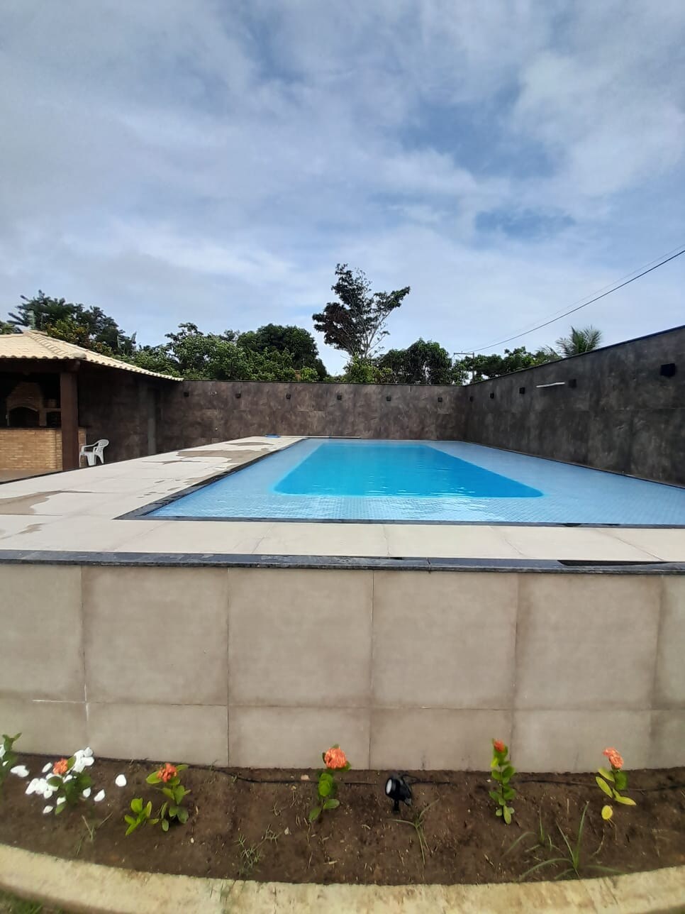 A spacious outdoor area is highlighted by a large pool with a clean, inviting surface, surrounded by a simple stone wall. Lush greenery and a few flowering plants are visible at the base, while an outdoor kitchen with a covered area is present in the background.