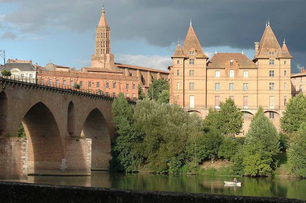 A historic stone bridge spans a river, leading to a view of elegant buildings characterized by red brick architecture. Towering trees with lush greenery line the banks, providing a natural backdrop. A small boat is gently gliding on the water, adding a serene element to the scene.
