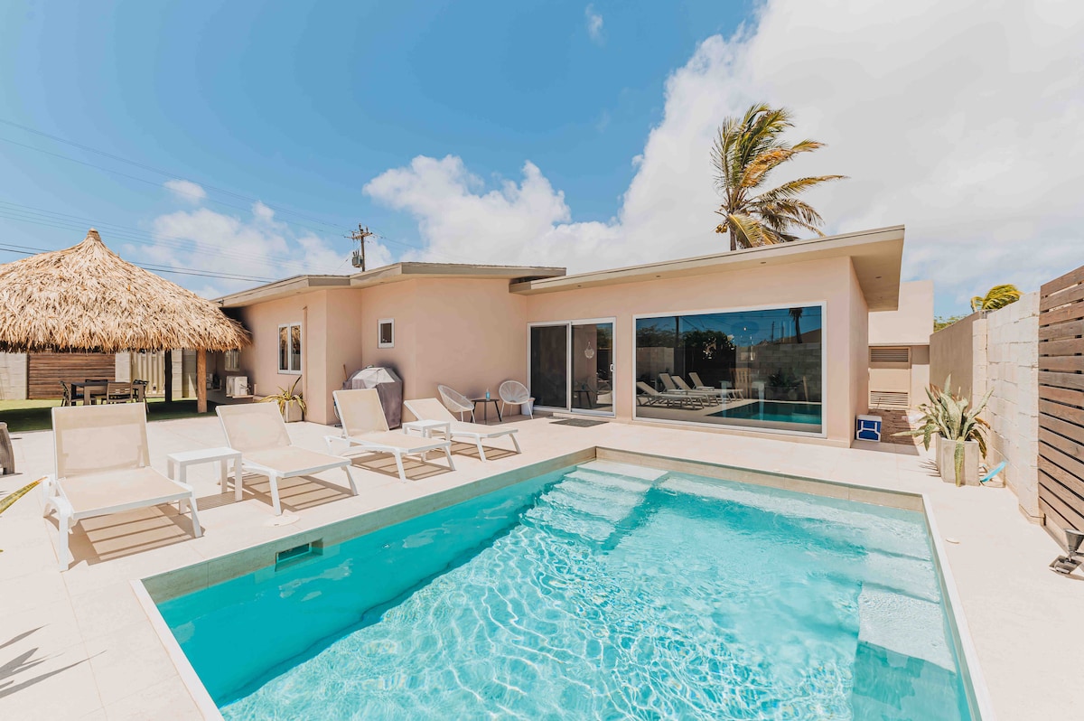 A serene outdoor area features a crystal-clear pool surrounded by sun loungers, with a shaded cabana nearby. The villa's large glass doors allow natural light to fill the interior, while palm trees sway gently in the breeze against a clear blue sky.